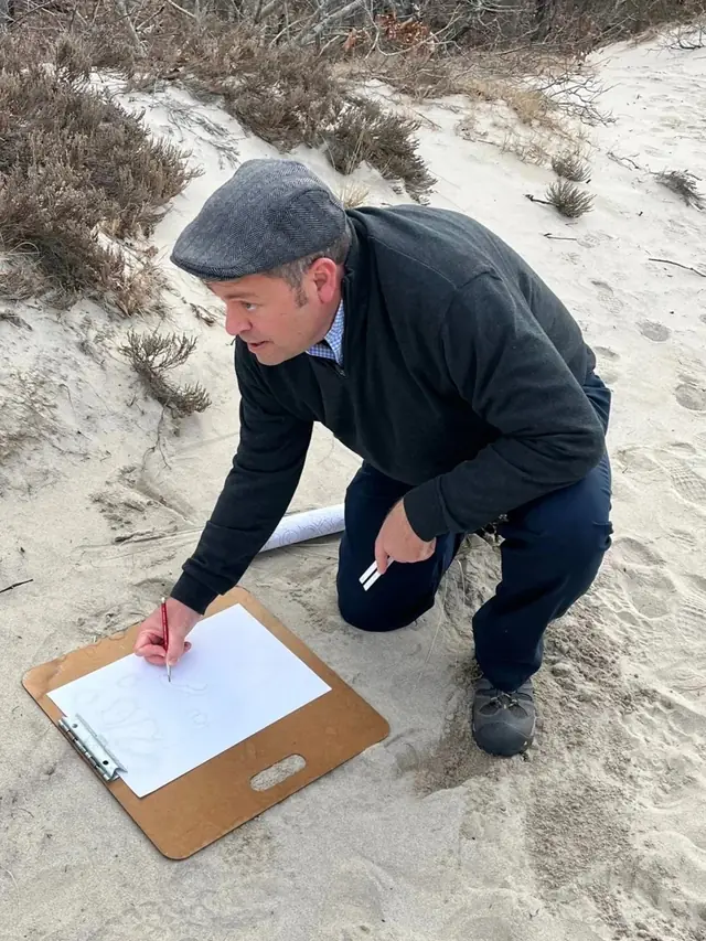 Golf course architect kneeling in coastal dunes, sketching bunker shapes on a clipboard in red pencil